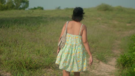 Woman-in-summer-dress-walking-through-grassy-field-at-dusk,-handheld-shot