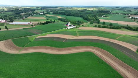 Aerial-view-of-curving-agricultural-fields-with-farm-buildings,-surrounded-by-greenery