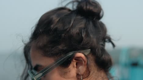 Close-up-of-a-young-woman-on-the-beach,-hand-in-hair,-with-sunglasses-reflecting-blue-skies,-contemplative-mood