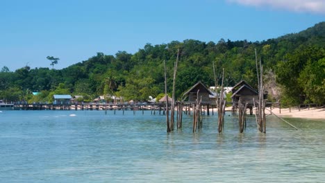 Slow-pan-of-scenic-exotic-tropical-island-with-white-sandy-beach,-wooden-beach-huts-overlooking-turquoise-ocean-water-on-rainforest-tree-covered-island-in-Raja-Ampat,-West-Papua,-Indonesia