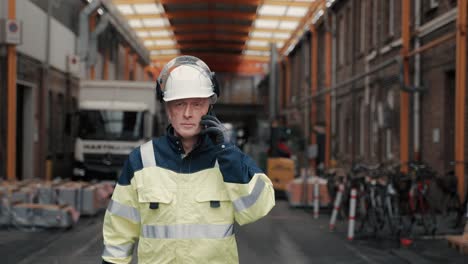 Caucasion-male-construction-worker-or-fireman-in-yellow-workwear-and-white-safety-helmet-at-the-phone-answering-call-walking-straight-to-the-camera-out-of-a-industrial-site-with-a-serious-worried-look