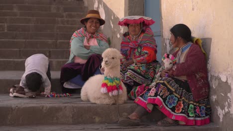 Indigene-Frauen-In-Cusco,-Cholitas,-Posieren-Fröhlich-Für-Ein-Foto-In-Ihrer-Traditionellen-Kleidung.-Die-Pollera-Neben-Ihnen-Ist-Ein-Alpaka,-Das-Strategisch-Platziert-Wurde,-Um-Touristen-Anzulocken,-Und-Ein-Kind-Ist-Beim-Spielen-Zu-Sehen