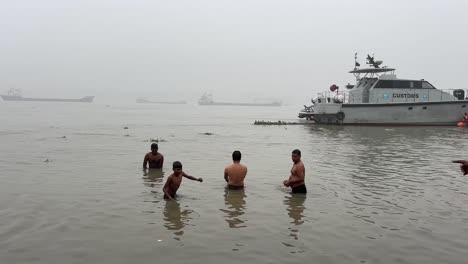 Vista-Trasera-De-Unos-Pocos-Hombres-Y-Niños-Bañándose-Y-Ofreciendo-Oraciones-Durante-Una-Tarde-Nublada-En-Babu-Ghat,-Kolkata.