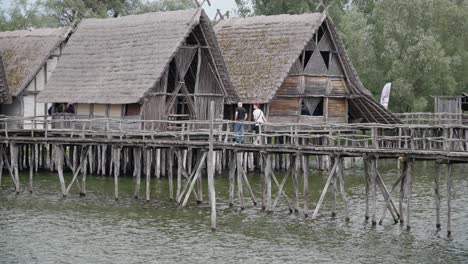 Thatched-roof-houses-on-stilts-over-water,-visitors-on-the-walkway,-historical-reconstruction,-Ice-Age-Museum-Friedrichshafen
