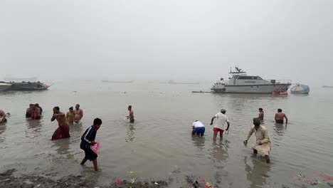 Men-and-woman-bathing-and-praying-in-foggy-winter-morning-during-Sankranti-with-jetty-in-the-background-during-a-cold-evening-in-Babughat,-Kolkata