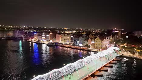 Handelskade-Willemstad-shining-lights-reflect-on-water-above-Queen-Emma-bridge,-aerial-descend