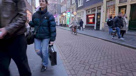 Amsterdam-thoroughfare,-diverse-pedestrians,-woman-pushing-a-stroller,-man-inspecting-bicycle,-shopping-bags,-winter-clothing,-narrow-street,-Dutch-buildings,-hanging-garlands,-chilly-ambiance