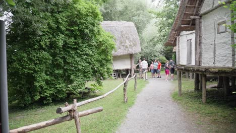 Group-of-people-touring-traditional-thatched-roof-cottages-at-an-Ice-Age-museum,-surrounded-by-lush-greenery