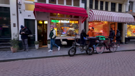 Urban-Amsterdam-street-scene,-bustling-sidewalk,-Pasta-Burger-restaurant,-awning,-people-conversing,-bicycles-parked,-overcast-weather,-festive-lights-strung-above,-European-architecture