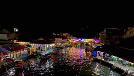 Night-view-of-a-vibrant-floating-market-with-illuminated-shops-and-moored-boats,-reflecting-colors-on-water