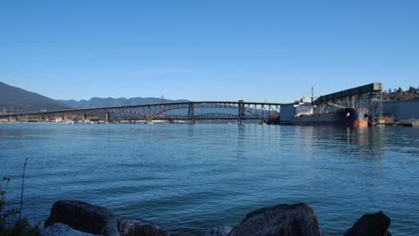 Calm-waters-with-a-view-of-Vancouver's-industrial-harbor-and-bridge-against-a-mountain-backdrop,-clear-day