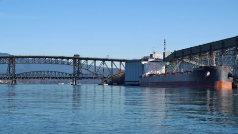 Frachtschiff-Im-Industriehafen-In-Vancouver-Angedockt,-Sonniger-Tag-Mit-Klarem-Blauen-Himmel,-Brücke-Im-Hintergrund,-Ruhiges-Wasser