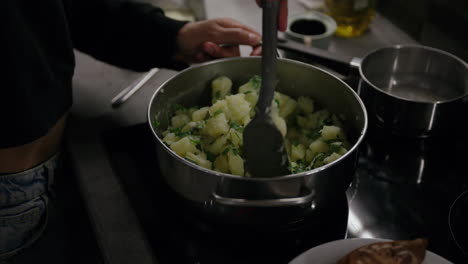 Close-up-shots-of-hands-mixing-potatoes-in-a-pot-on-the-stove-using-a-turner,-woman-cooking-dinner-in-the-kitchen,-crushed-potatoes-with-parsley,-European-side-dish,-vegetarian-food,-vegan-diet,-pot