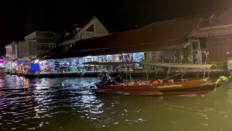 Nighttime-view-of-a-vibrant-floating-market-with-illuminated-signage,-shops,-and-a-moored-boat-on-a-lively-river