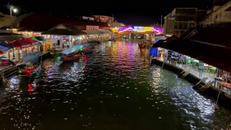 Animado-Mercado-Nocturno-Junto-Al-Río-Con-Coloridos-Puestos-Y-Barcos,-Luces-Vibrantes-Que-Se-Reflejan-En-El-Agua