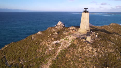 A-solitary-lighthouse-on-a-rugged-coastline-with-ocean-backdrop,-clear-sky,-aerial-view