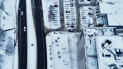 Drone-top-down-snow-covered-parking-lot-as-cars-exit-onto-highway,-satellite-view