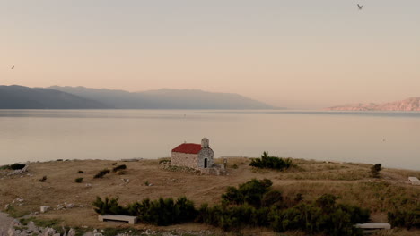 Cinematic-drone-shot-revealing-a-tiny-island-with-a-church-in-the-middle-of-the-sea,-sunrise,-blue-hour,-seagulls-flying-around,-calm-water,-peaceful-clear-view,-chapel,-Novi-Vinodolski,-Croatia