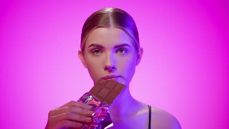 Blonde-nordic-girl-eats-a-chocolate-bar-suggestively-in-a-studio-with-pink-background,-close-up-portrait-shot