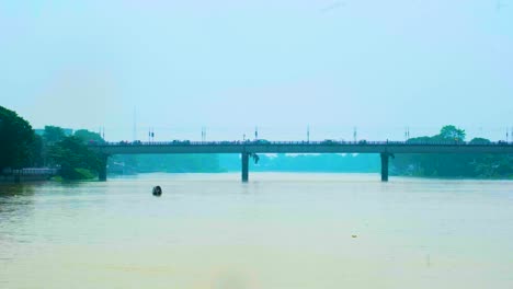 Un-Pequeño-Barco-En-Un-Ancho-Río-Bajo-Un-Puente-En-Medio-Del-Tráfico-Sobre-él-En-Bangladesh,-Sylhet