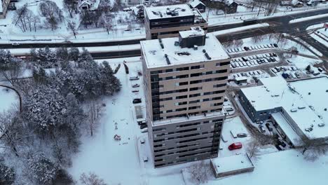 Modern-grey-beige-apartment-building-covered-in-snow-next-to-coniferous-forest,-drone