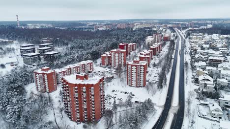 Brown-walled-high-rise-buildings-covered-in-snow-as-cars-drive-below-on-highway,-aerial-overview
