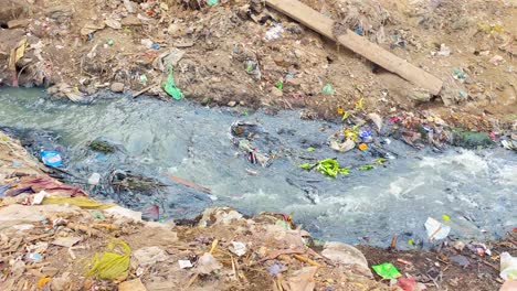 Las-Aguas-Residuales-Salen-Disparadas-De-Una-Tubería-Rodeada-De-Basura-Y-Fluyen-A-Lo-Largo-De-Un-Banco-De-Barro-Sucio,-Pasando-Por-Un-Perro-Carroñero,-Hacia-Un-Río-En-Bangladesh.