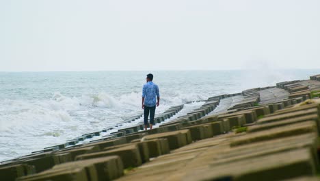 Lonely-Man-walking-along-a-sea-defense-as-waves-crash,-loneliness-concept