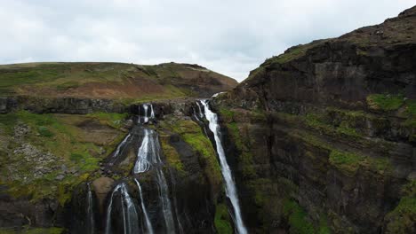 Water-drapes-over-volcanic-rocks-covered-with-green-moss-on-Glymur-waterfalls,-aerial-dolly
