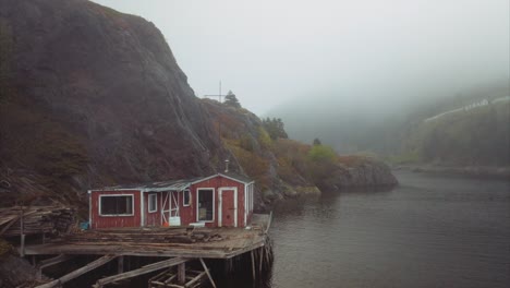 Uninhabited-wooden-home-on-stilts-over-calm-water-nestled-against-steep-cliffs-with-foggy-background