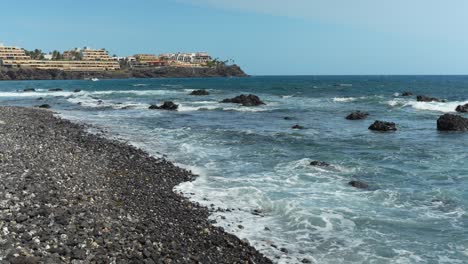 Small-ocean-waves-gently-crash-on-pebble-rocky-beach-with-beachside-hotels-in-background