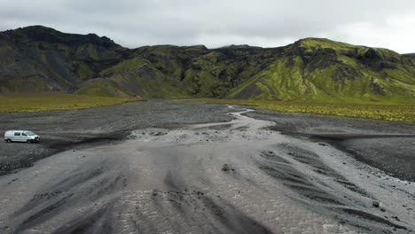 White-van-driving-over-solid-magma-shallow-river-flowing-from-Icelandic-volcanic-mountain-range