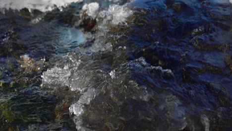 Water-flows-over-rocks,-snow-and-ice-in-shallow-stream-bed,-closeup