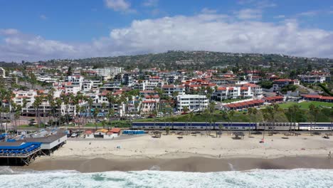 4k-Aerial-Video,-left-to-right-showing-train-passing-San-Clemente-Pier-and-beach-and-shops-in-Orange-County,-towards-San-Diego,-California,