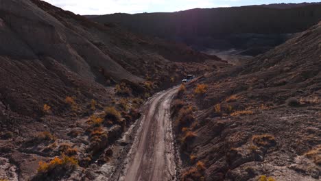 Descending-aerial-white-off-road-SUV-driving-on-dirt-road-through-desert-landscape
