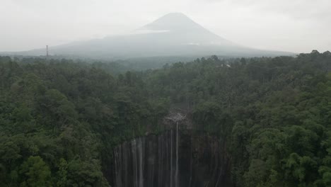Aerial:-Tumpak-Sewu-waterfall-in-Java-jungle,-volcano-cone-in-distance
