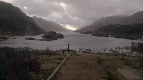 Glenfinnan-monument-by-the-lake-with-scottish-highlands-in-backdrop,-overcast-sky,-aerial-view