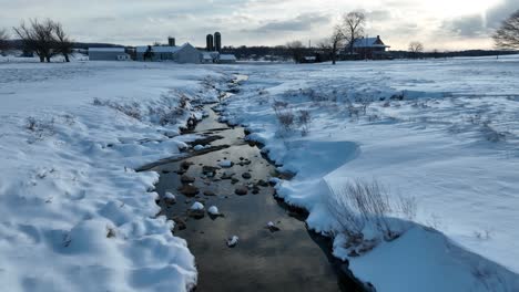Snowy-landscape-with-a-meandering-stream-near-a-farm-under-a-cloudy-sky
