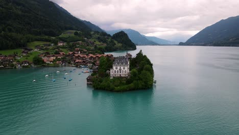 Iseltwald-castle-on-a-peninsula-in-a-swiss-lake,-with-a-backdrop-of-a-quaint-village-and-mountains,-aerial-view