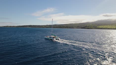 Catamaran-drives-across-open-ocean-with-tropical-cloudy-covered-mountains-on-coast,-aerial-orbit