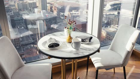 a-small-table-set-in-the-kitchen-of-a-luxury-high-rise-condo-next-to-large-glass-windows-showing-the-city-beneath-it