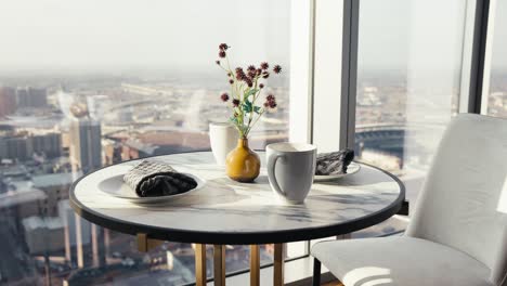 a-small-table-set-with-flowers-coffee-cups-and-plates-next-to-large-windows-in-a-downtown-high-rise-condo