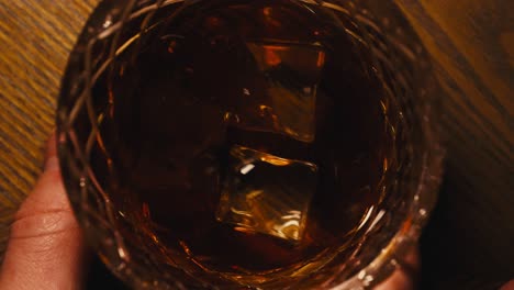 Nahaufnahme-Von-Oben-Nach-Unten-In-Zeitlupe-Eines-Mannes,-Der-Ein-Glas-Bourbon-Whisky-Auf-Einem-Holztisch-Schwenkt