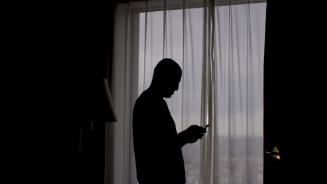 Silhouette-of-a-man-standing-near-the-window-with-the-curtains-closed,-typing-on-a-mobile-phone-screen,-the-concept-of-privacy-and-anonymity