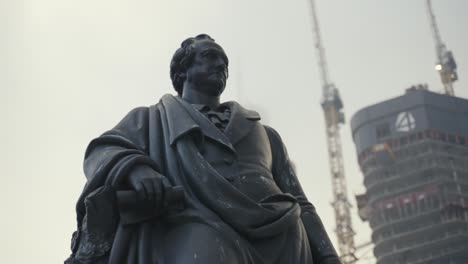 Close-up-of-the-statue-of-goethe-with-tower-four-skyscraper-in-the-background-in-Frankfurt-germany-on-a-foggy-cold-winter-day-while-at-goethe-platz-while-the-tower-is-under-construction