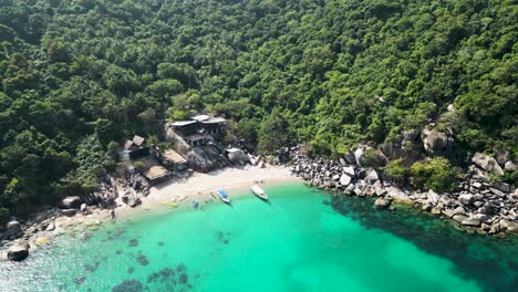 Seaside-cabins-with-boats-on-an-inlet-in-Mango-Bay-Ko-Tao-Island-Thailand-with-tourists-at-beach,-Aerial-descent-approach-shot