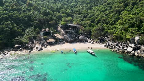 Boats-on-an-inlet-with-cabins-in-Mango-Bay-Ko-Tao-Island-Thailand-with-tourists-at-beach,-Aerial-descent-tilt-up-shot