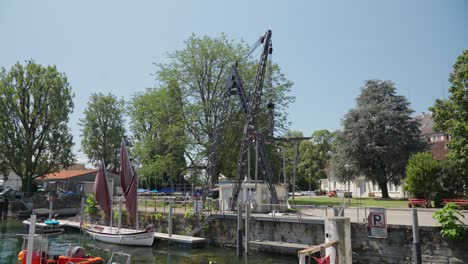 Traditional-sailboat-docked-by-a-calm-river-with-a-crane-and-greenery-in-Lindau,-Bodensee-on-a-sunny-day