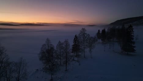 Snowy-landscape-at-dusk-with-fog-over-the-fields-and-a-colorful-sky-near-sundsvall,-sweden,-aerial-view
