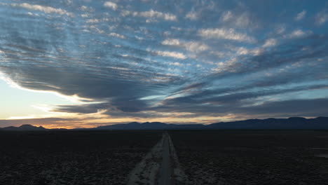 Desert-road-stretching-into-horizon-under-a-vast-sky-with-dramatic-clouds-at-dusk,-Mojave-Desert,-timelapse